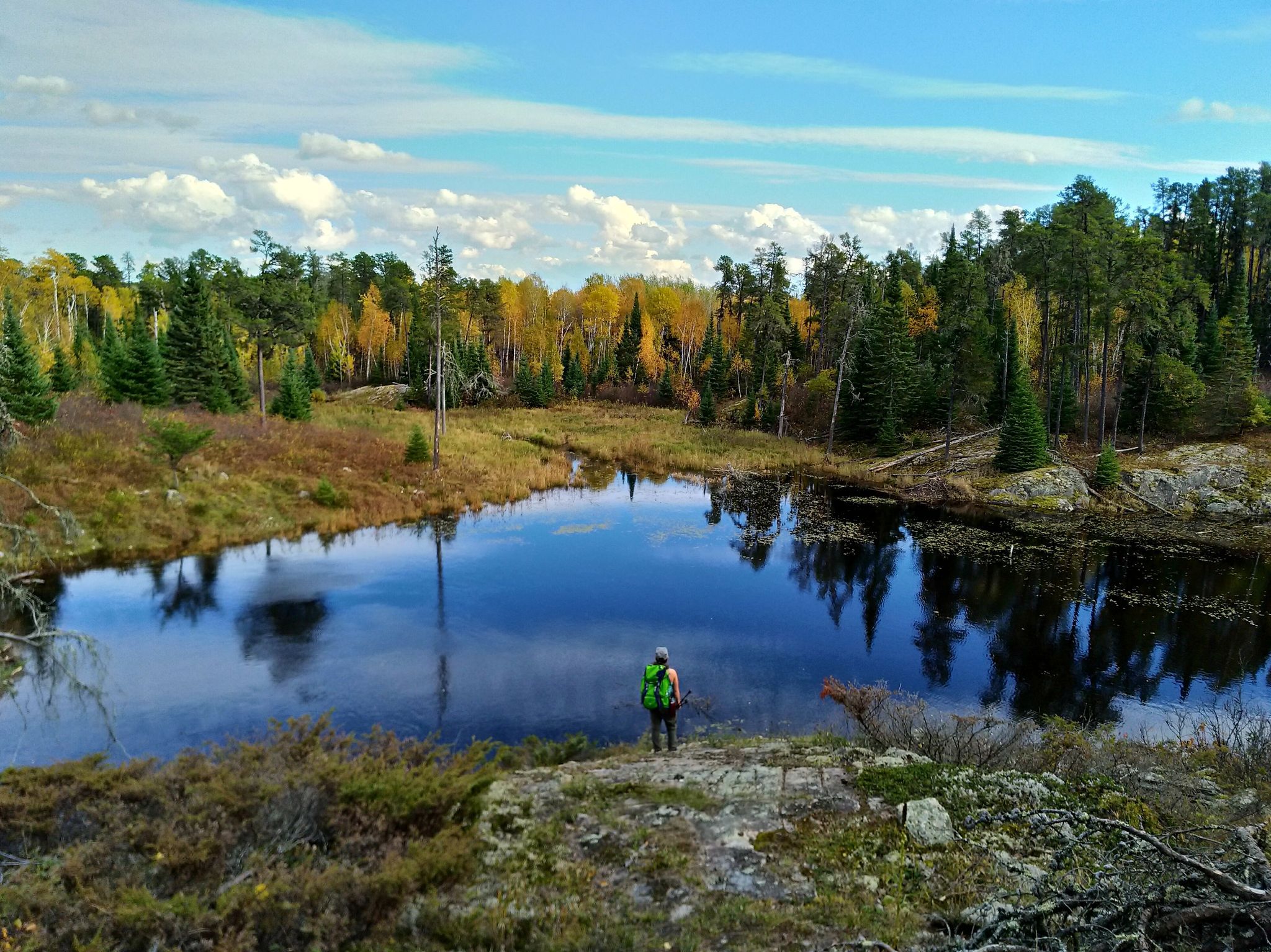 a hiker stands in front of a glassy lake surrounded in boreal forest and rocky banks