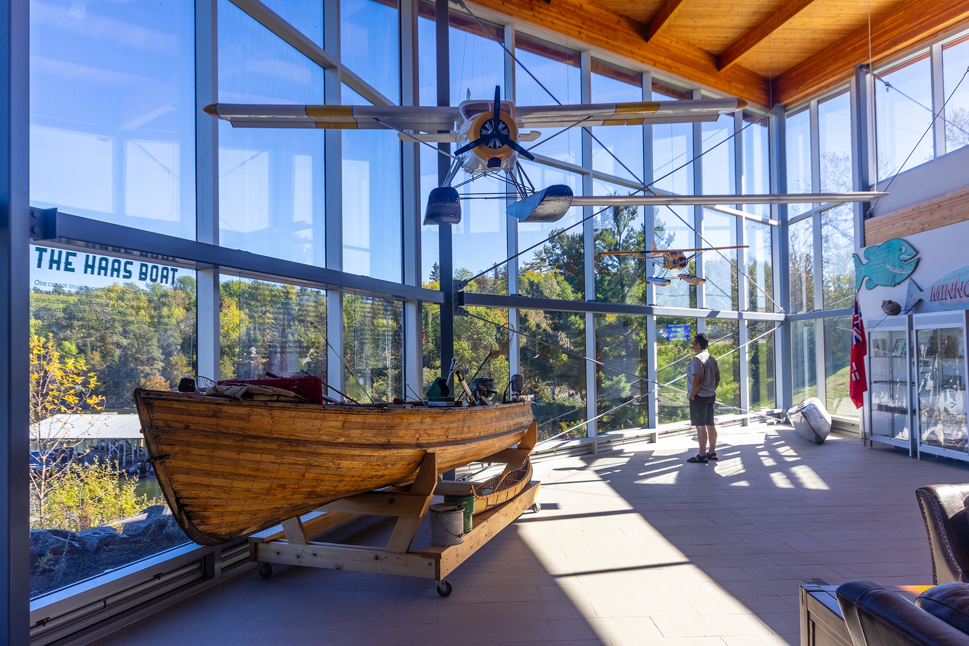 A man looks out of the windows in a large sunlit atrium next to an antique wooden boat and a small float plane.