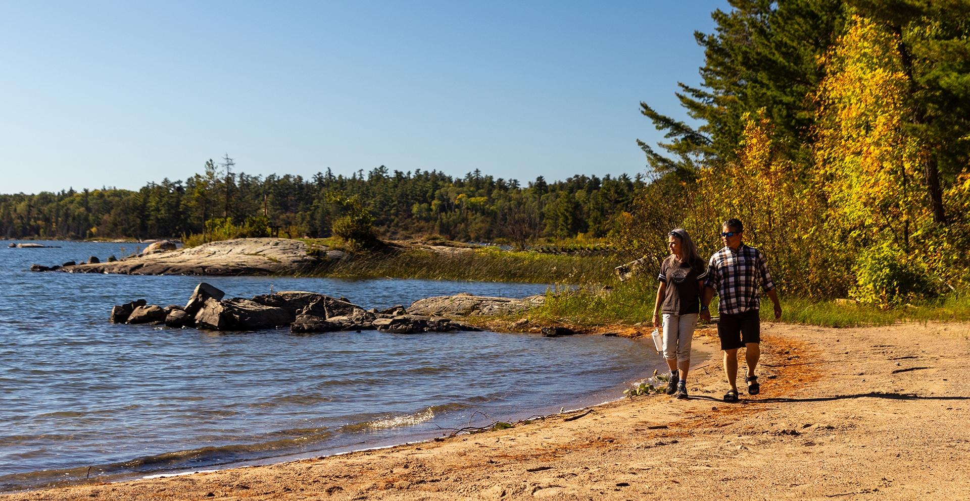 Two people walk along a sandy beach beside a blue lake