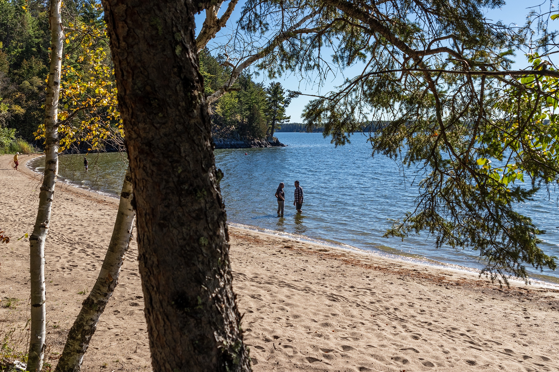 Two people wading in the lake beside a long sandy beach