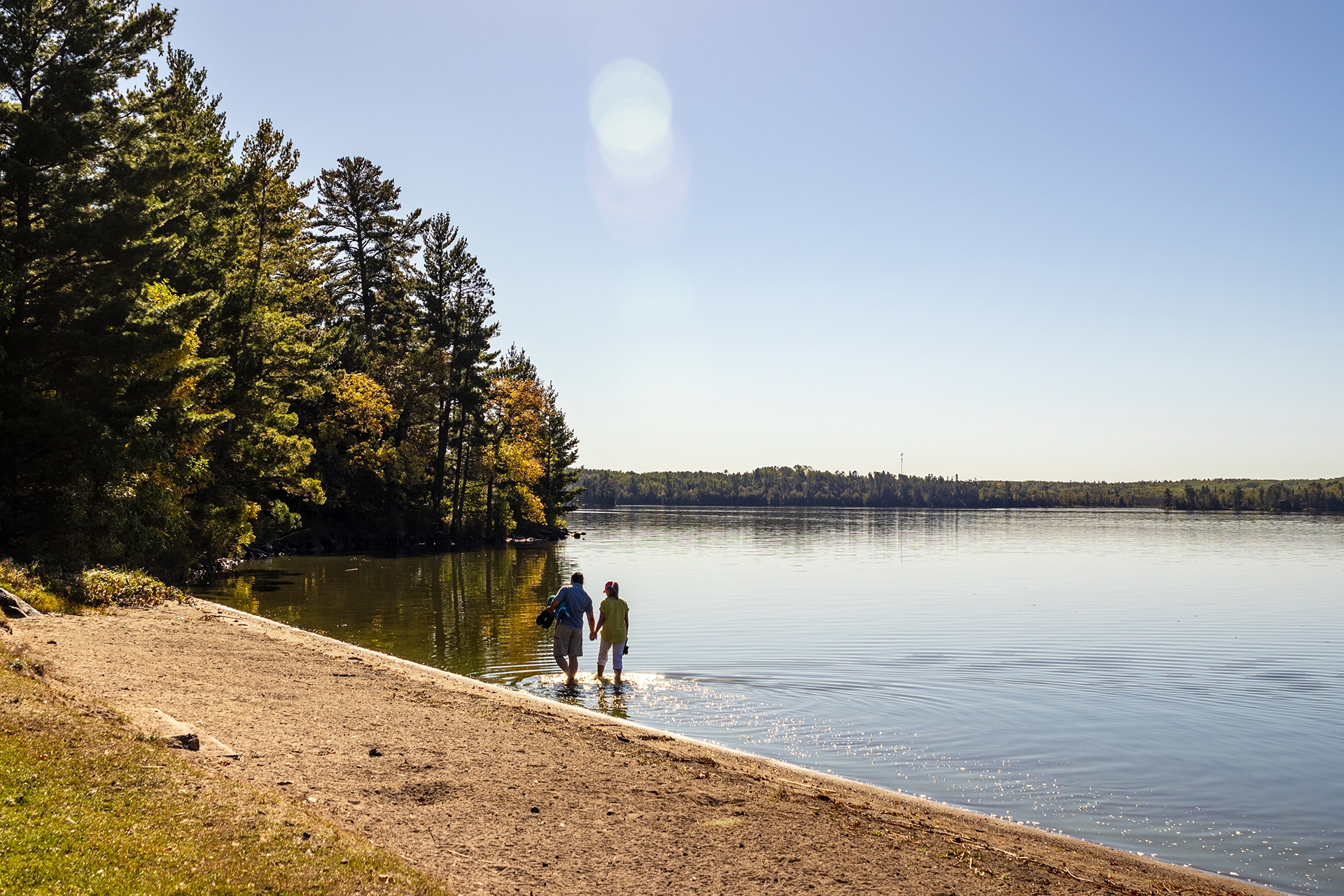 Two people hold hands and wade through water beside a sandy beach