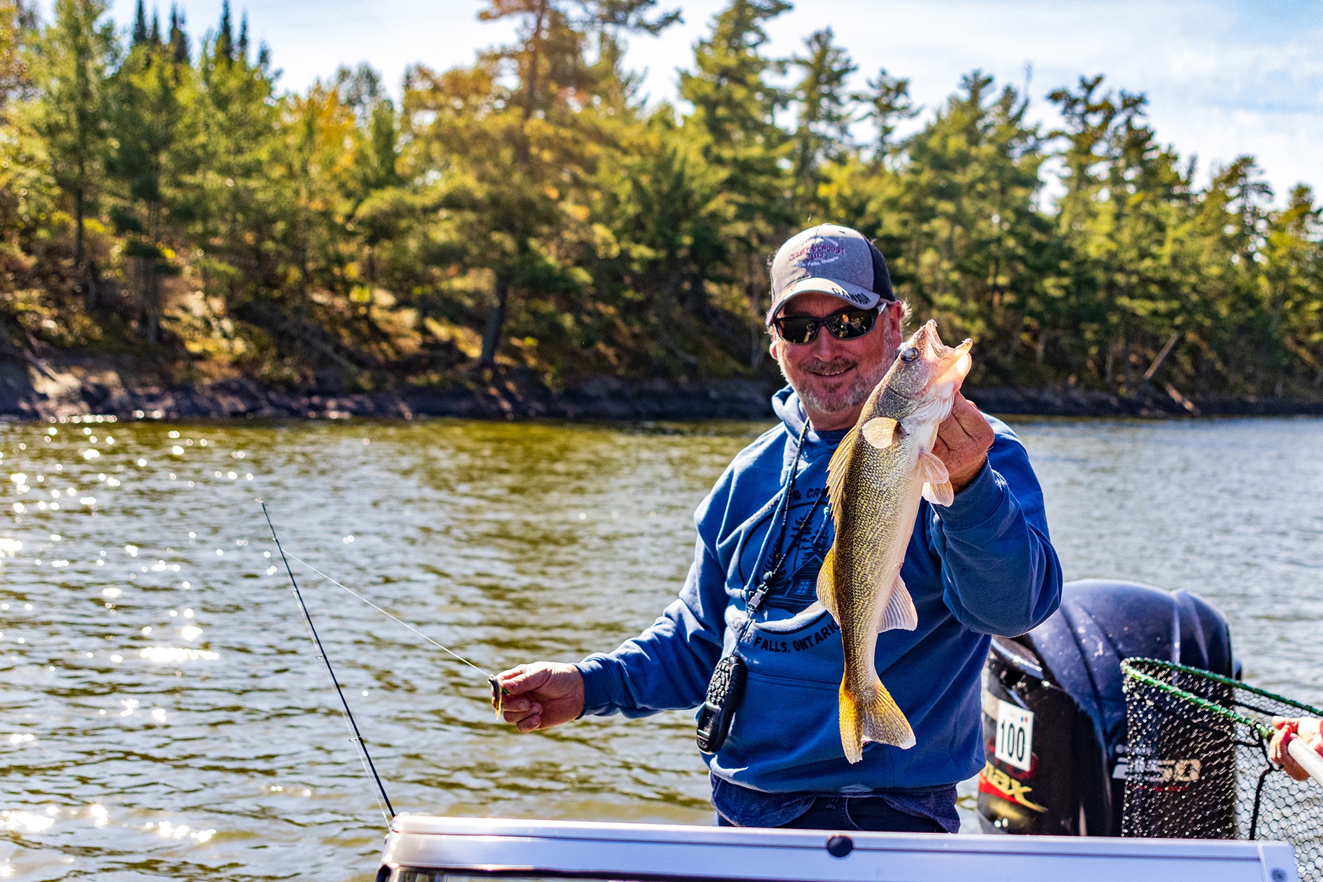 A man wearing sunglasses and a baseball cap holds up a large fish he's just caught