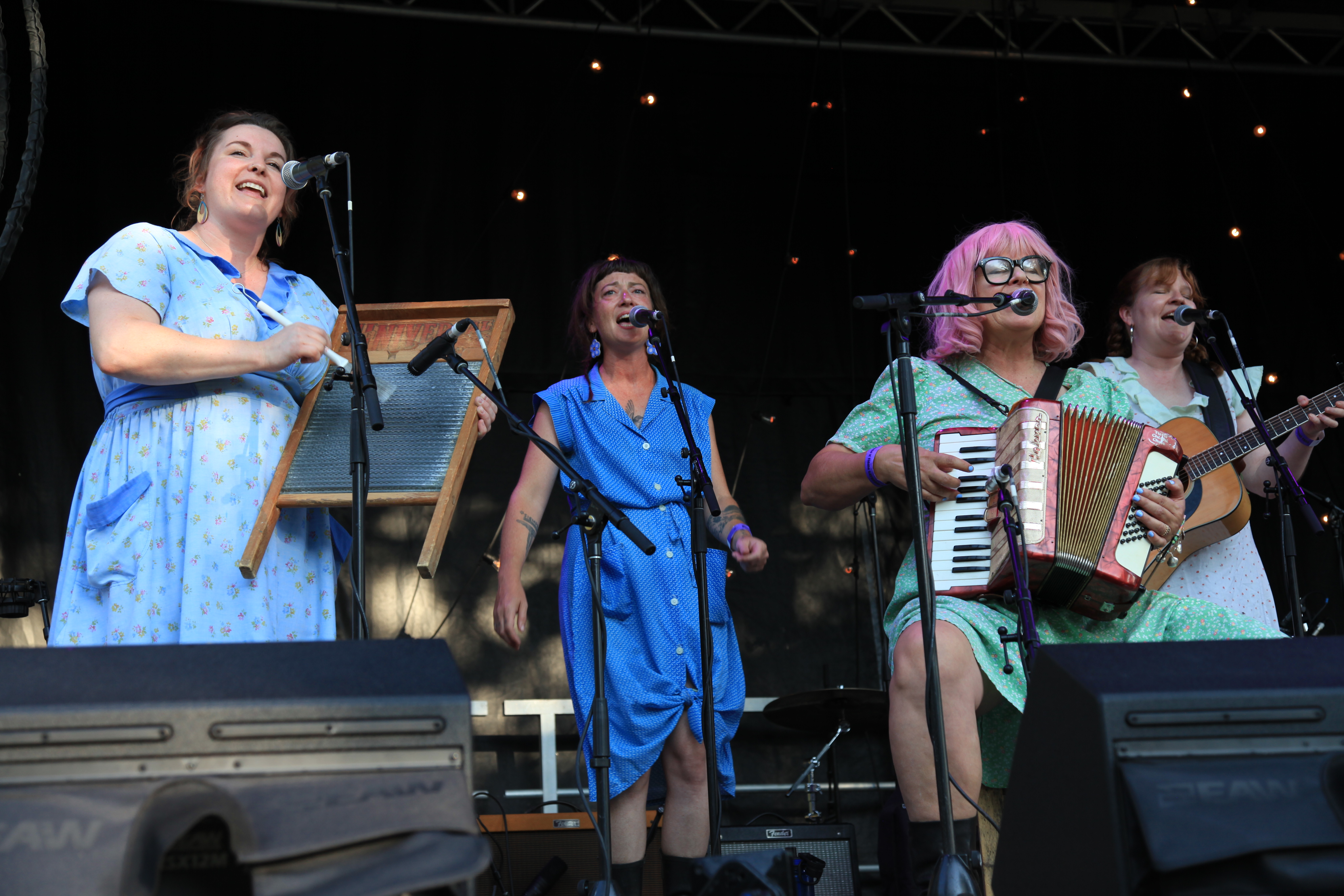 Four women in colourful dresses sing into microphones and play folk instruments