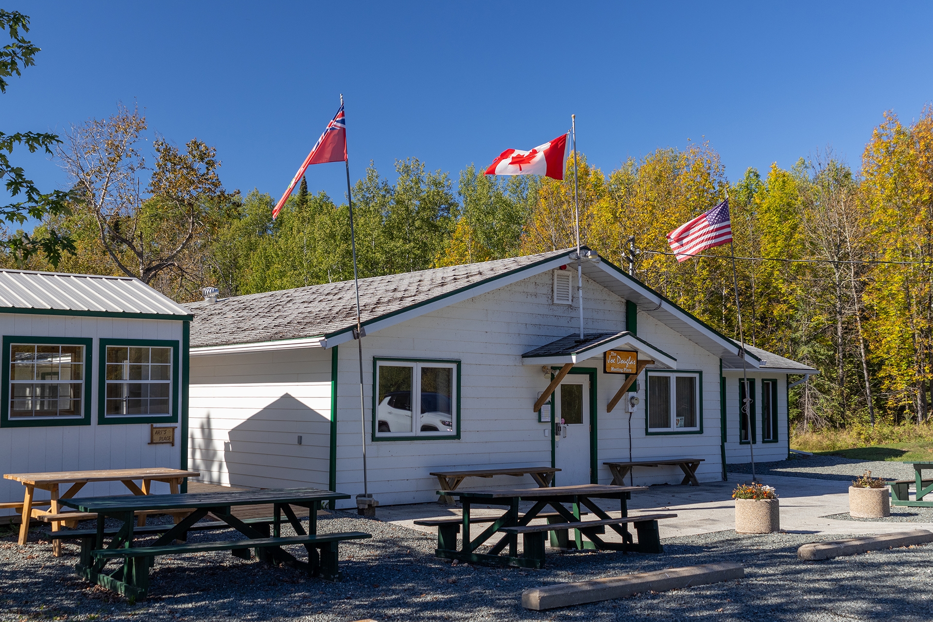a modest white building in front of green forest with wooden picnic tables out front, Canadian and American flags flying overhead and a small sign that reads "The Joe Douglas Meeting Place"